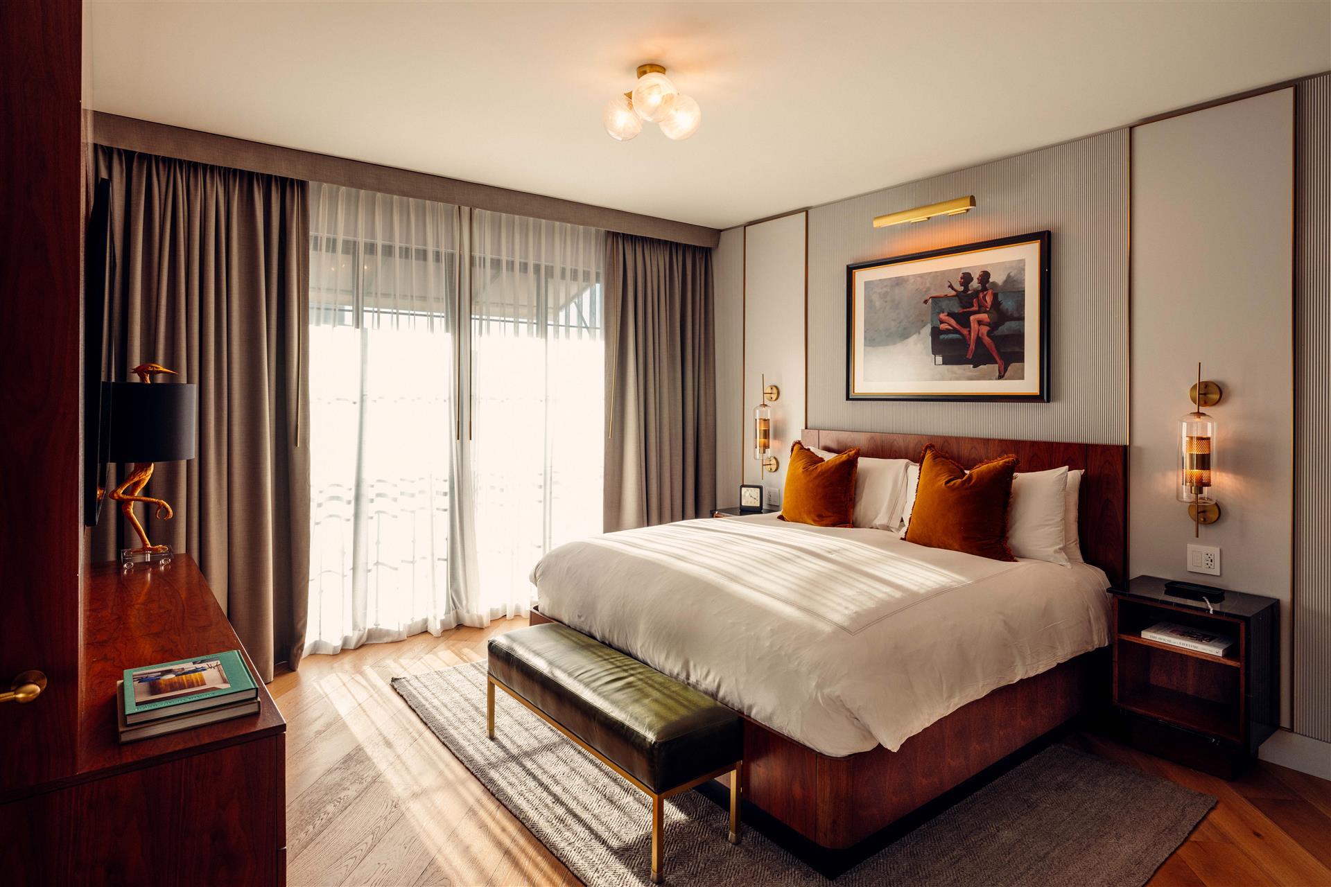 A sunlit bedroom with a wood headboard, rust-colored cushions, an olive bench, brass lights and framed artwork.